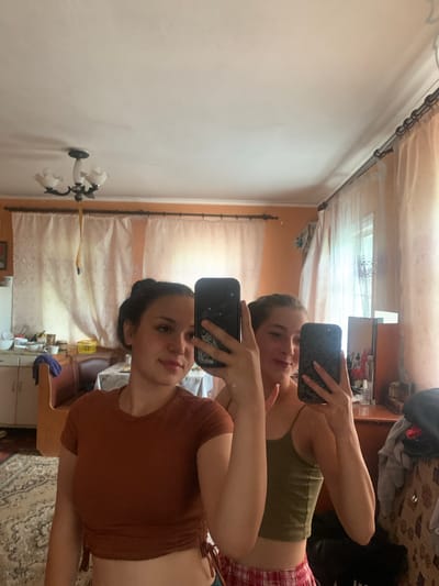 The image is a vertically oriented selfie taken in a room, likely a bedroom, as indicated by the presence of a bed frame and curtains. Two young women are visible, reflected in a mirror. One woman in the foreground is wearing a brown, cropped t-shirt and her hair is pulled back. She is holding a phone in her right hand, taking the picture. The second woman, slightly further back, is wearing a green tank top and plaid shorts. She is also holding a phone, taking her own selfie. Both women appear to be looking at their phones. The room's decor is simple, with light-colored walls and patterned curtains. There are some objects visible on a surface behind the women, including what appears to be a shelf with bottles and containers, suggesting a dresser or vanity. The lighting suggests it is daytime. The image is a candid moment, capturing the two friends taking selfies in what appears to be a casual setting. There is no visible text in the image.