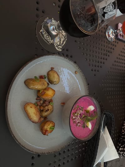 An overhead shot captures a dimly lit table setting featuring a plate of roasted potatoes and a bowl of vibrant pink soup, likely served in a restaurant. The potatoes, some halved, are scattered on a textured grey plate, garnished with diced bacon and chopped green onions. The soup, a bright pinkish-purple hue, is in a clear glass bowl and topped with sprouts, dill, and slices of hard-boiled egg. A dark glass, possibly containing red wine or a dark beverage, rests on a black coaster with a goat logo and text. A second, smaller glass with a red liquid is visible further back. The table surface is dark and perforated, suggesting an outdoor or industrial-style setting. A black woven basket, a white napkin, and cutlery are also present on the table. The overall ambiance suggests a casual dining experience.