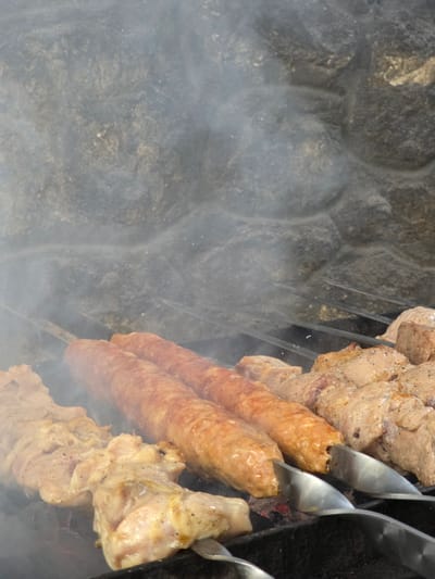 This is a close-up, eye-level shot of various types of meat being grilled on skewers over charcoal. The meat appears to be a mix of chicken pieces and long, cylindrical sausage-like kebabs. Smoke rises from the grilling meat, obscuring the background which consists of a rough, grey stone wall. The scene is likely outdoors, possibly in a backyard or park area in Pidhorodne, Ukraine, given the rustic nature of the stone and the popular barbecue activity. There are no people visible in the image. The focus is entirely on the food and the grilling process.