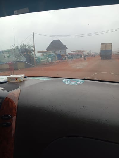 The image shows a view from inside a vehicle, looking through the windshield at a street scene in Imilike, Nigeria. The road is dirt and appears to be a main thoroughfare with several buildings on either side. A gas station with multiple pumps is visible on the left, and a large truck is seen driving away on the right. There are also several people and motorcycles on the street, suggesting a bustling urban environment. The sky is overcast, and the overall atmosphere is hazy. A Swiss air freshener is placed on the dashboard of the vehicle in the foreground.