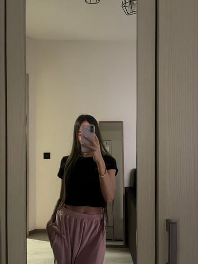 A female individual is depicted in an indoor setting, reflected in a large mirror. The exact location within Novosaratovka, Russia, cannot be determined from the image. The individual, whose face is obscured by a light purple smartphone with a patterned case, is wearing a black short-sleeved crop top revealing her abdomen and light pink sweatpants. Her long, dark hair extends over her shoulders and partially covers her chest. Her left hand is placed in her pants pocket, while her right hand holds the smartphone, positioned to capture her reflection. A red string bracelet with a white bead is visible on her right wrist. The reflected environment features light-colored walls and flooring, with two black geometric pendant light fixtures visible on the ceiling. A black rectangular light switch is affixed to the wall on the left side of the reflection. Further into the background, another rectangular mirror with a wooden frame is visible, reflecting a portion of the room, alongside a dark piece of furniture. The main mirror appears to be part of a door or closet, indicated by a handle on its right side and a vertical textured panel adjacent to it.
