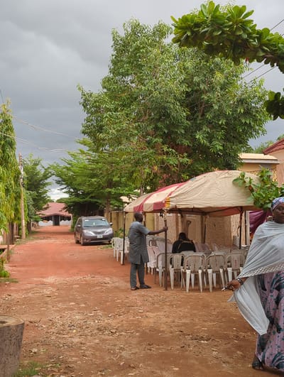 The image depicts an outdoor scene in Unguwar Rimi, Nigeria, characterized by reddish-brown dirt ground and an overcast, grey sky. Lush green trees, with varied foliage, dominate the left side of the frame, with power lines visible extending across the top and mid-ground. In the background, buildings with light-colored walls and reddish-brown roofs are partially visible.
On the right side, a temporary canopy or awning, beige with red stripes, is set up, supported by poles. Beneath it, numerous white plastic chairs are arranged and stacked, indicating preparations for an event or gathering. The word "RENTALS" is clearly visible printed on the backs of several chairs, suggesting they were hired for the occasion.
Several individuals are present. On the far right, a woman wearing a light-colored head covering (hijab) and a patterned dress stands, looking towards the camera. She holds a dark object, possibly a pen, in her hand. Closer to the center, under the canopy, a man in a dark grey suit or uniform is standing, holding an upright pole, possibly involved in the setup or organization of the event. Other figures are partially discernible under the canopy and among the chairs in the background.
A dark-colored minivan or car is parked on the dirt path in the mid-ground. A large, cut log or tree trunk is partially visible in the bottom right corner. The overall scene suggests a community gathering or outdoor event taking place during the daytime, under cloudy weather conditions.