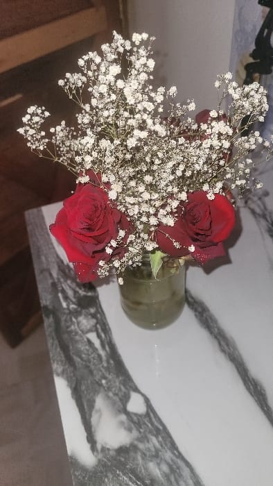 The image, taken in Nanyuki, Kenya, depicts a close-up, slightly elevated perspective of a floral arrangement situated on a polished surface. The central elements are three deep red roses, exhibiting closed to partially open petals and some green stem foliage, interspersed within a substantial cluster of small white flowers, identified as Baby's Breath (*Gypsophila*). These flowers are arranged within a transparent glass jar, which contains a clear liquid filling approximately one-quarter of its volume. The jar rests on a flat, reflective surface characterized by a white base with prominent irregular grey and black veining, consistent with marble or a similar decorative stone. The background features a blurred, dark brown, possibly wooden structure or wall segment in the upper left. To the mid-right, a light grey, out-of-focus wall is visible, with a dark, indistinct object or fixture partially discernible at the extreme right edge of the frame. No individuals, actions, or interactions are present in the scene.