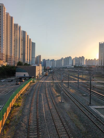 The image shows a high-angle view of train tracks curving through an urban landscape. The scene is set in Daegu, South Korea. A line of tall apartment buildings runs along the left side of the frame, their surfaces bathed in the warm light of the setting or rising sun. Adjacent to the buildings is a parking lot with several parked cars. A green barrier separates the parking lot from the train tracks. The tracks, which are multiple and parallel, stretch across the right side of the frame and curve into the distance. Overhead electrical lines and support structures run above the tracks. The ground between and around the tracks is covered in gray gravel. In the distance, more buildings are visible, gradually fading into the hazy sky. The overall lighting suggests either sunrise or sunset.