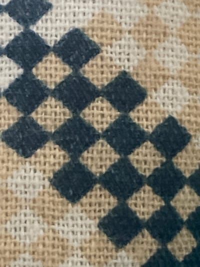 The image shows a close-up view of a woven fabric with a diamond pattern. The fabric appears to be made of natural fibers, with alternating threads of a light tan and a white or off-white color. Overlaid on this base weave are dark blue diamond shapes. These diamonds are not perfectly formed, with slightly uneven edges and a textured appearance, suggesting they are embroidered or a printed pattern on the fabric. The lighting is even, and there are no shadows that indicate a specific time of day or environment beyond the fabric itself. The image does not contain any elements that would allow for the identification of a specific location within the city of Ардонь, Russia.