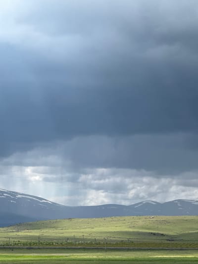 The image depicts a landscape near Tlik, Armenia. The right side of the frame is dominated by a green, grassy field with utility poles running parallel to the edge. Adjacent to the field are mountains with patches of snow. The sky takes up the majority of the frame, ranging from a dark blue and cloudy region to lighter, cloudier area with apparent sun rays.