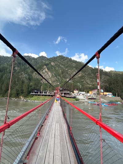 A person wearing a red swimsuit and blue shorts walks away from the camera on a wooden suspension bridge. The bridge has red railings and metal mesh netting. The person is carrying a yellow plastic bag in their right hand. The bridge spans a wide, murky river. In the background, on the far bank, is a complex of buildings with multi-colored roofs, including a prominent dark building with multiple levels and balconies. Beyond the buildings, a steep, forested mountain rises under a bright blue sky with scattered white clouds. The area appears to be a tourist destination, with several inflatable rafts or boats visible on the river near the buildings. The scene is likely in Barangol, Russia, given the presence of a suspension bridge over a river and the type of architecture suggestive of a mountain resort area.