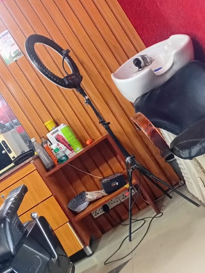 The image displays the interior of a salon located in Dorayi, Nigeria. A black ring light on a black tripod stand occupies the central foreground, positioned vertically with its light ring illuminated. To the upper right, a white ceramic hair washing basin with a chrome faucet is mounted against a red-painted wall. Below the basin, a black salon chair with a distressed, cream-colored seat is partially visible.
To the left, a wooden cabinet features both drawers and open shelves. The top shelf holds multiple bottles, including a green one labeled "W MARK" and a yellow-capped bottle, alongside an orange spherical object. The lower shelf contains a black electronic device, a folded piece of paper, and a black comb-like object. Below these shelves, two multi-socket electrical outlets are mounted on the wall. The power cord from the ring light extends across the light-tiled floor towards these outlets.
In the lower-left foreground, a portion of another black salon chair with chrome armrests is visible. The wall behind the ring light and cabinet is constructed from vertical wooden panels, with a white poster displaying text and a scooter image partially visible on the left side. No individuals are present within the scene.