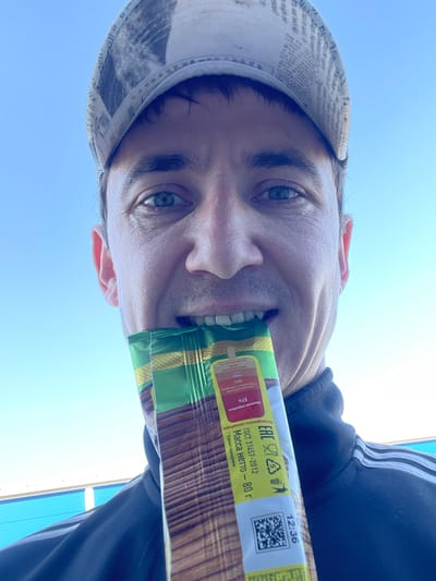 A man, wearing a patterned baseball cap and a black jacket, is shown from the chest up, holding a food bar in his mouth, with the packaging facing the camera. The background consists of a clear blue sky with a faint horizontal blue and white structure in the distance, likely a building or fence. The food bar's packaging features Russian text and logos. The scene appears to be outdoors.