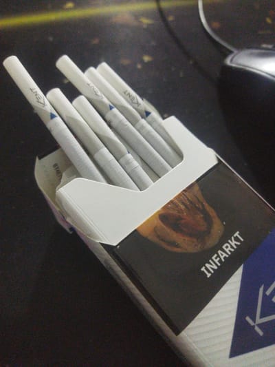 The image shows a pack of cigarettes, specifically "INFARKT" brand, with "KENT" written on the filter of several cigarettes. The pack is open, revealing the cigarettes arranged inside. The background appears to be a dark, textured surface, possibly a table or desk, with some out-of-focus elements including what looks like the edge of a computer mouse. The lighting is dim, creating some graininess in the image. The scene depicts a still life of cigarettes. There is no indication of the city of Zafar, Uzbekistan, or any elements that would place it there.
