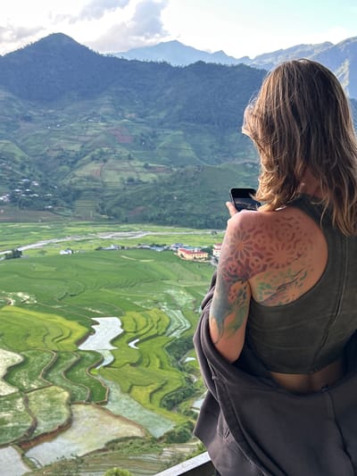 An elevated view captures a person from behind, looking out over the terraced rice fields of Tú Lệ, Vietnam. The individual has medium-length brown hair and is wearing an olive green tank top, with a dark grey zippered hoodie draped over their shoulders. A prominent, multi-colored tattoo featuring geometric and organic patterns is visible on their right shoulder and upper arm. The person holds a black smartphone in their right hand, its screen reflecting the overcast sky. Below, lush green rice paddies, some filled with water reflecting the sky, cascade down the valley, following the contours of the land. A light-colored river or stream winds through the valley floor, with scattered buildings and small villages nestled among the fields. In the distance, multiple layers of verdant, forested mountains rise under a cloudy sky, with hints of blue sky overhead. A metal railing or ledge is partially visible in the lower right foreground, indicating an observation point.