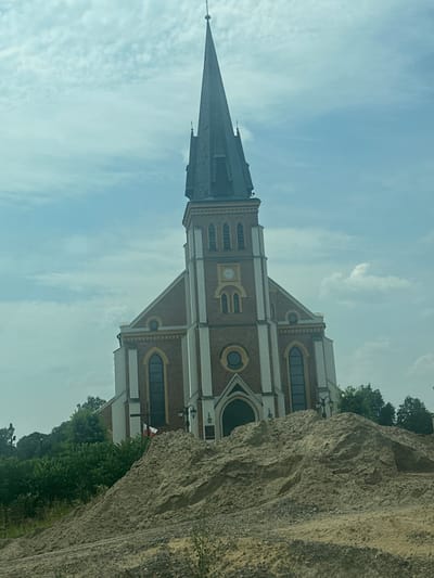This is a landscape orientation media file, rotated 90 degrees counter-clockwise, depicting a church with a tall, pointed steeple. The church appears to be constructed of red brick with white trim around arched windows and decorative elements. A clock is visible on the church facade. To the right of the church, a large rock formation dominates the foreground, obscuring part of the building and surrounding trees. The sky above is cloudy with a light blue hue, suggesting daytime. The overall scene is of a church and its natural surroundings, with the rock formation adding a dramatic element. There is no visible text in the image. The location is Nienadówka, Poland.