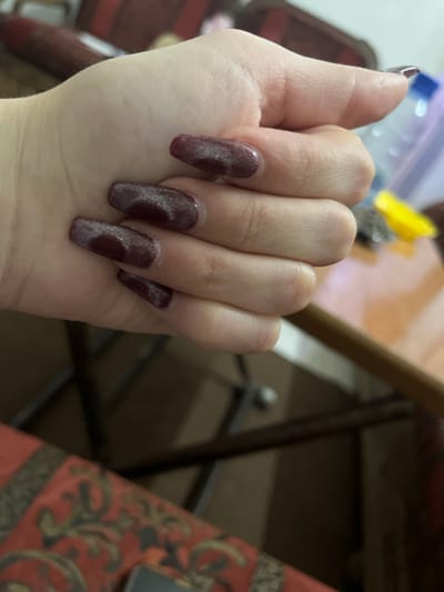 A left hand, palm towards the camera, displays manicured nails. The nail polish is a deep maroon color with silver shimmers and a cloudy pattern. The nails are rectangular and appear freshly done. The skin of the hand is pale. Background objects include a clear plastic bag, yellow and silver objects, a wooden table edge, a brown metal folding chair, and a red patterned tablecloth.