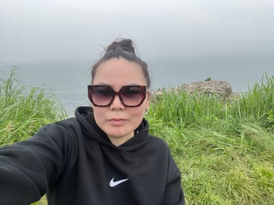 A woman takes a selfie outdoors on a foggy day. She is wearing a black Nike hoodie and large, tortoise-shell sunglasses. Her hair is pulled up in a bun. Behind her, a body of water, likely the Sea of Okhotsk, is visible through a thick fog. In the middle ground, a rocky outcrop covered in birds can be seen. The foreground is dominated by tall green grass. The location is Tikhoe, Russia.