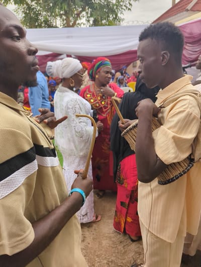 This image depicts an outdoor gathering in Unguwar Rimi, Nigeria.
In the left foreground, an adult male with a dark complexion is seen in profile, facing right. He has short dark hair and a light beard. He wears a tan polo shirt with a horizontal black and white stripe across the chest. His left hand holds a small, light-colored hand drum, and his right hand holds a curved wooden beater. A blue wristband is on his right wrist.
In the right foreground, a second adult male with a dark complexion is also in profile, facing left, towards the first man. He wears a light yellow, vertically striped long-sleeved shirt with a matching fabric draped over his left shoulder and light-colored trousers. He holds a small, light-colored hand drum against his body with his right arm and hand, and a curved wooden beater in his left hand. He has short dark hair.
In the midground, between the two men, an adult female with a lighter complexion is visible in profile, facing right. She wears a white headwrap, a white lace dress, gold hoop earrings, and eyeglasses. To her right, another adult female wears a red headwrap and a red dress with patterned details. The background contains additional individuals, predominantly women in various colorful traditional garments and headwraps, some standing and some seated. A yellow plastic chair is partially visible behind the woman in red.
The background features white and maroon fabric drapes, indicating a temporary canopy or tent structure. Above the drapes on the right, barbed wire fencing is visible. Green tree foliage is present at the top of the frame. The ground is a light-colored, textured surface. The scene is illuminated by diffused natural light, consistent with daytime.