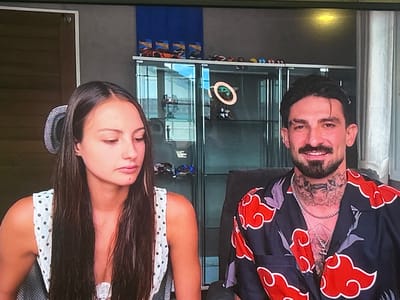 The image shows a medium shot of a man and a woman sitting next to each other. The woman is on the left, with long dark hair and wearing a white dress with black polka dots. She is looking to her left. The man is on the right, with short dark hair, a mustache, and a beard. He is wearing a black shirt with red cloud patterns and has multiple tattoos on his neck and chest. He is smiling and looking at the camera. In the background, there is a glass display cabinet with toys and collectibles on the shelves. The overall environment appears to be an indoor setting, possibly a living room or office. There is no indication of the city of Boboshevo, Bulgaria, in the image.