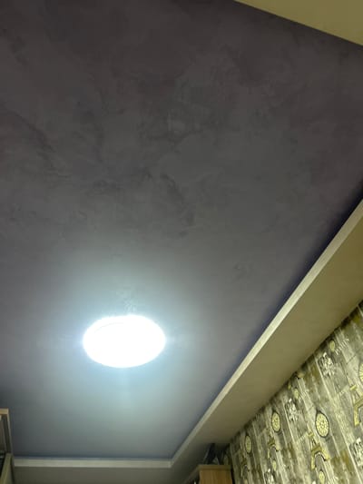 A low-angle interior shot shows a textured, blue-violet wall with a bright, circular light fixture. The ceiling has a patterned wallpaper featuring images of bicycles and the Eiffel Tower. The scene is illuminated by the light fixture and an unspecified light source. A beige-colored trim separates the wall and ceiling. Parts of a cabinet or furniture are visible in the lower right corner. There's no indication of the image's location being Voluyak, Bulgaria.