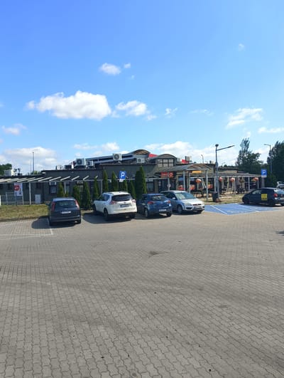 The image depicts a sunny day at a service station in Połupin, Poland. The foreground is dominated by a gray interlocking paver parking lot. Several cars are parked in the lot, including a dark gray Ford Focus with license plate FKR 338CX, a white Nissan X-Trail with license plate FZ 33921, a blue Mazda 2 with license plate PRW 3J7E5, and a silver Ford Fiesta with license plate POZ 1V231. A disabled parking spot, marked with blue lines and a wheelchair symbol, is also visible.
In the background, a modern building with a dark gray facade houses a Circle K convenience store and what appears to be a fast-food establishment with outdoor seating. The building features large windows, an overhang supported by pillars, and a prominent red and white Circle K logo. Several outdoor air conditioning units are mounted on the roof. A tall lamppost stands in front of the building. The sky is a clear blue with scattered white clouds. The overall scene is one of a typical roadside service area.