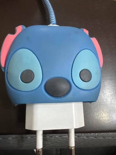 A close-up, head-on shot shows a blue, Stitch-shaped phone charger adapter. The adapter has large, light blue oval eyes with dark grey pupils, a black nose, and pink triangular ears. A blue, corrugated cord extends from the top of the adapter. The adapter is plugged into a white iPhone power brick, which is sitting on a dark, textured surface that appears to be leather or faux leather. The lighting is direct and casts minimal shadows. There are no people or discernible actions taking place in the image. The scene is devoid of any contextual information relating to Yeghegnadzor, Armenia, as the image is solely focused on the charger adapter and its immediate surroundings.
