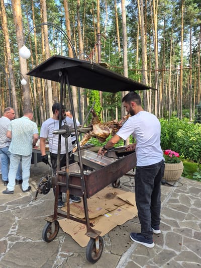 In Zelyonovka, Russia, a group of men are gathered around a large rotisserie grill in an outdoor setting. The grill is positioned on a cobblestone patio, surrounded by tall, slender pine trees and lush green foliage. A whole lamb is slowly roasting on a spit above the open fire. One man, dressed in a white chef's coat and black pants, is actively tending to the lamb, using tongs to adjust its position. He wears black gloves. Two other men stand nearby, observing the cooking process. One man wears a light-colored, patterned shirt and light pants, while another wears a white striped shirt and jeans. A fourth man, also in a chef's attire, stands behind the rotisserie. The scene suggests a catered event or outdoor feast taking place in a natural, wooded environment.