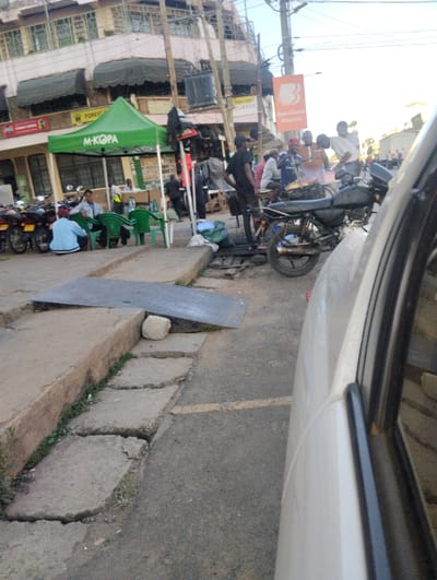 The image captures a vibrant street scene in Sheywe ward, Kenya, bathed in bright daylight under a clear sky. In the foreground, the side of a white car is visible on the right, overlooking a partially paved road and a dilapidated concrete sidewalk. A crude metal ramp bridges an exposed drain or ditch on the sidewalk, held in place by a large stone. The sidewalk exhibits cracks and small patches of grass.
On the left side of the street, a prominent green tent with the "M-KOPA" logo is set up, indicating a mobile money or service point. Under the tent, several individuals are seated on green plastic chairs, some interacting with a person behind a small table. A row of motorcycles, commonly used as boda-bodas, are parked nearby. Behind the tent, buildings with green awnings and a visible "Coca-Cola" sign line the street.
Further down the street, a lively gathering of men is centered around what appears to be a street food stall, with one person tending to food on a grill or hot plate. A black motorcycle is parked near this group. Overhead, a large orange sign for "Bank of Baroda" is visible, featuring text in both English and likely Gujarati or Hindi script. Another banner above displays the word "TUSKER," a local brand. The buildings in the background are multi-storied, and numerous power lines crisscross the upper part of the frame, characteristic of an active urban commercial area.