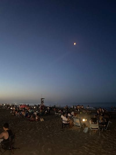This is a vertical, wide-angle, nighttime photograph taken from a low perspective on a beach. The sky is a deep gradient of indigo to a lighter blue, with a hazy horizon suggesting the presence of lights from a distant town or city. A bright, circular object, possibly the moon or a lantern, is visible in the upper left portion of the sky.
The foreground and right side of the image are dominated by a sandy beach crowded with people. Many are seated in folding chairs, facing the water, which is visible in the far left of the frame. The scene appears to be an outdoor gathering or event, possibly a concert or a fireworks display, given the large number of people and the anticipation suggested by their posture. Some individuals are standing, and the overall atmosphere seems relaxed and social.
Objects visible include numerous beach chairs, some with striped fabric, and a few beach umbrellas. A lifeguard stand with a person visible at the top is situated on the beach, indicating this is a designated swimming area. Several portable lights are illuminating parts of the beach, casting a warm glow.
The time of day is clearly night, transitioning from dusk into darkness. The weather appears to be clear, with no visible precipitation. The location cues, such as the beach setting, the presence of a lifeguard stand, and the distant city lights, align with the provided context of Canet d'en Berenguer, Spain. There is no visible text in the image.
