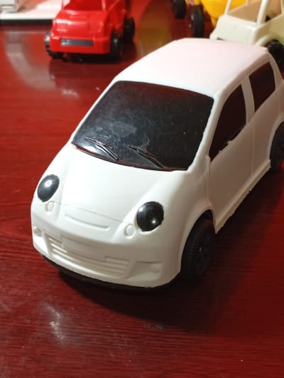 A white toy car is positioned on a red, reflective surface. The car has black windows and black wheels. In the background, a red toy car and a yellow toy vehicle are partially visible. The red surface appears to be a table or desk. The lighting suggests an indoor environment. The image does not contain information about the city of Khujayli, Uzbekistan.