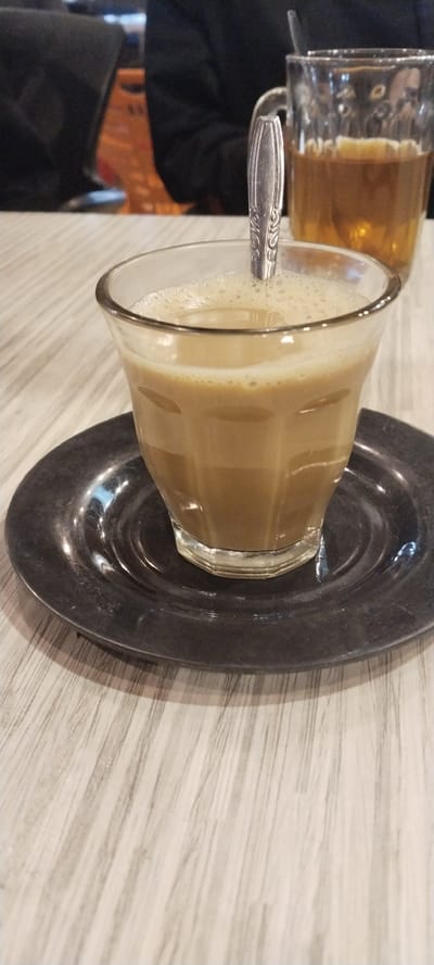 A close-up, eye-level shot shows a glass cup filled with a frothy, light-brown beverage, likely coffee or tea, resting on a dark, metallic saucer. A silver spoon with intricate detailing is partially submerged in the drink, its handle extending upwards. The cup and saucer are placed on a light-colored wooden table with a visible grain pattern. In the background, slightly out of focus, is another clear glass mug containing a darker amber liquid, possibly tea or coffee. Behind the mugs, a person wearing a dark garment is seated, with part of their shoulder and torso visible. Further in the background, a hint of orange and black fabric is visible, possibly a bag or clothing item. The overall setting appears to be indoors, possibly a cafe or restaurant. The scene captures a moment of simple enjoyment of beverages.
