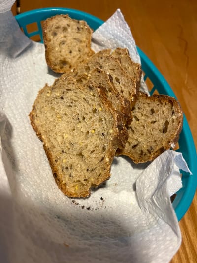 A close-up, elevated shot shows several slices of artisanal bread resting in a teal-colored plastic basket, lined with a white paper towel. The bread appears to be a multi-grain or sourdough variety, characterized by its rustic, dark crust and a visible crumb with small holes and embedded seeds, possibly including flax and yellow mustard seeds or millet. There are at least five slices visible, cut relatively thick. Some dark crumbs or specks are present on the white paper towel inside the basket, particularly towards the right side. The background consists of a warm-toned wooden surface, likely a table, suggesting an indoor setting. The lighting is soft and ambient. The image depicts a simple presentation of bread, possibly for serving at a meal, in Chêne-Bourg, Switzerland. No people or text are visible.
