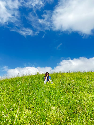 A young woman with dark hair and glasses is sitting in a field of tall green grass under a bright blue sky with scattered white clouds. She is wearing a blue shirt and white pants. The grass is dense and appears to be growing on a gentle slope. Small yellow flowers are visible interspersed within the grass. The woman is looking to her right and resting her chin on her hand, appearing contemplative. The overall scene depicts a peaceful moment in a natural, outdoor setting. The image does not contain enough information to definitively place the location in the city of Среднее Кечево, Russia.