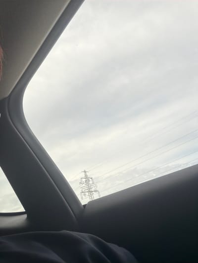 The image captures a view from inside a vehicle, looking out through the windows. In the foreground, the dark interior of the car is visible, including a portion of what appears to be a person's shoulder and clothing. Through the side window, the overcast sky dominates the frame, with several power lines stretching horizontally across the scene. On the right side of the window, a large electrical pylon is visible, with text and symbols printed on the glass in front of it. The overall lighting suggests a cloudy or dimly lit day. The scene does not provide any specific visual cues to indicate it is in Vallensbæk Landsby, Denmark, or any other specific city.
