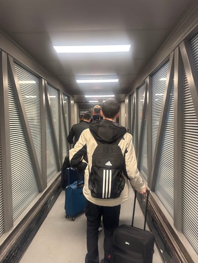 An image, originally oriented vertically but presented here rotated 90 degrees, captures the interior of an airport jet bridge, likely located at Geneva Airport (GVA) in Le Grand-Saconnex, Switzerland. The scene depicts several travelers moving away from the viewer, suggesting they are either boarding or disembarking an aircraft.
The primary subject, seen from behind in the foreground, is a male wearing a light-colored, possibly beige or off-white, jacket with dark accents. He is carrying a black backpack featuring a distinctive white Adidas logo and three parallel white stripes. With his right hand, he pulls a black rolling suitcase with an extended handle. Another blue rolling suitcase is visible on the floor closer to the foreground subject's feet. Further down the jet bridge, additional individuals and luggage are visible, receding into the distance.
The jet bridge itself is characterized by its modern, functional design. The left wall features several bright, vertical fluorescent light panels set against a plain grey surface, providing ample illumination. The ceiling and right-side wall are constructed with corrugated metal panels, supported by dark grey structural beams, creating a repetitive pattern and industrial aesthetic. The floor is a light-colored, possibly reflective, surface. The internal lighting suggests a well-lit indoor environment, making the external time of day or weather conditions indiscernible. The overall atmosphere conveys a typical airport travel experience of transit and purposeful movement.