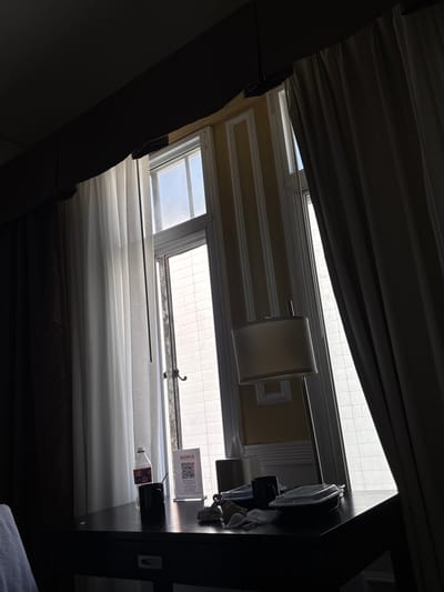 A dimly lit room has a view of a window with sheer curtains partially drawn to the left and heavy curtains to the right. The window is divided into multiple panes, and through it, an overcast sky and the exterior of a building with light-colored brickwork are visible. On a dark table in front of the window, a partially empty bottle of Coca-Cola is standing upright, next to a black mug. To the right of the mug is a white sign with a QR code and text that reads "SOMA". A floor lamp with a cream-colored shade stands behind the table, to the right of the Coca-Cola bottle. The room appears to be a hotel room, as suggested by the style of the window and the presence of a hotel information sign. The scene is captured from a low angle, looking upwards towards the window. The overall impression is one of a quiet, possibly early morning, setting in a hotel room in San Francisco, California.