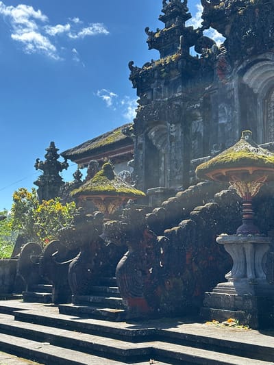 The image shows a portion of the Pura Besakih temple complex, specifically a stone structure with moss and intricate carvings. The structure is in Rendang, Indonesia, and the sky is visible with some clouds. There are some plants visible at the bottom right corner of the image.