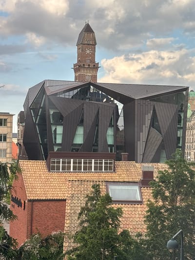 This media file is an outdoor shot of a modern building with a unique geometric facade, likely a commercial or public building, in Malmö, Sweden. The building features a dark, angular exterior with large glass windows. To the left of the frame, a tall, traditional brick tower with a pointed roof stands against a sky filled with dramatic, light-colored clouds. The sky suggests it is daytime, possibly late afternoon or early evening given the lighting. The scene is framed by green trees on the right side of the image, partially obscuring another building with a tan tiled roof and a brick section. The overall impression is a juxtaposition of historical architecture with contemporary design, set within an urban environment. There is no visible text in the image.