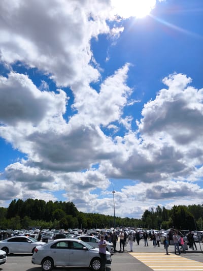 This media file depicts a busy outdoor parking lot, likely at Khrabrovo Airport in Russia, on a bright and partly cloudy day. The scene is dominated by a large expanse of paved asphalt filled with numerous cars and a multitude of people, with a backdrop of dense green trees.

In the foreground and midground, many individuals, including adults and some children, are visible. They are dressed in casual attire, moving about, walking across a distinctive yellow and white striped pedestrian crossing, or standing near parked vehicles. Their activity suggests arrivals, departures, or transit to and from a destination.

The parking lot is extensively populated with various makes and models of cars, primarily sedans and hatchbacks, with a noticeable prevalence of white and silver vehicles. These cars are either parked in marked spaces or slowly navigating the lot. A tall, slender utility pole stands prominently in the mid-distance, extending towards the sky.

The upper portion of the image is filled with a bright blue sky adorned with large, voluminous cumulus clouds. The sun is intensely bright in the upper right quadrant, producing a clear lens flare, indicating that the time of day is midday or early afternoon. The weather appears to be fair, with a mix of sunshine and cloud cover. The overall atmosphere suggests a bustling public area or transport hub. No discernible text is visible on any objects or signs.