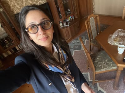 A person with dark hair and round glasses takes a selfie. They are wearing a navy blazer and a patterned scarf. In the background, a wooden table with a cup and plastic-wrapped item sits near a chair with floral upholstery. A wooden cabinet filled with items is partially visible, along with a bookshelf displaying books. The floor is covered with a patterned carpet. The picture is rotated slightly.