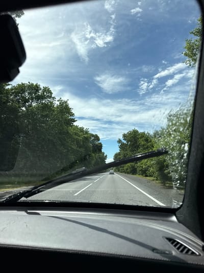 The image depicts a view from inside a vehicle driving on a paved road lined with dense green trees. The sky is blue with scattered white clouds. A white car is visible in the distance ahead on the road. The vehicle's windshield wipers are partially visible in the foreground. The dashboard of the vehicle is also visible at the bottom of the frame. The scene appears to be a daytime shot on a road in a rural or semi-rural area. There is no indication of the specific location within the city of Nizhnyaya Saniba, Russia.