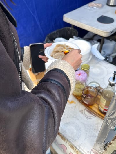 A person in a brown jacket with white fur trim is holding a plate of dumplings and a smartphone. The dumplings appear to be garnished with a sauce. Above the plate are bottles and jars of sauces and condiments arranged on a table. The table itself features a patterned white surface. A blue tarp is visible in the lower left corner.