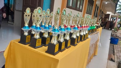 In Blangriek, Indonesia, a long table draped with a yellow cloth is set up outdoors, likely in front of a building. The table is laden with numerous golden trophies, each with a black base and a golden inscription plate. The trophies are arranged in neat rows, varying in color at the top, with blue, red, and green accents visible. Some trophies have circular emblems at their apex. In the background, individuals in uniforms, possibly students or staff, are seen standing and sitting near a building with windows. The environment appears to be a school or institutional setting, with a tiled floor, a walkway, and some vegetation visible to the right. A blue bucket and some building materials are also present on the ground. The lighting suggests it is daytime, with diffused light indicating overcast conditions or shade.