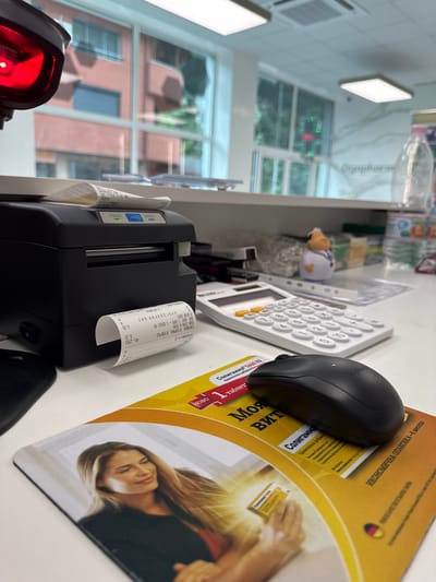 This is a close-up, angled shot of a retail counter in what appears to be a pharmacy or drugstore in Madan, Bulgaria. The dominant objects are a thermal receipt printer with a receipt partially dispensed and a black computer mouse resting on a promotional mousepad. The mousepad features a woman's face and text in Cyrillic script, possibly advertising a product. Behind the mouse and calculator, shelves with various items are visible, out of focus.

The receipt printer is actively in use, evidenced by the paper sticking out. The visible text on the receipt includes transaction details such as "ПРОДАЖБА № 31" (Sale No. 31), currency amounts in Euros and Bulgarian Leva ("ОБЩА СУМА ЕВРО," "ОБЩА СУМА В ЛЕВА"), and an exchange rate ("ОБМ. КУРС 1 ЕВРО 1.95583"). The promotional mousepad also contains Cyrillic text, including "ИКОНОМИЧНА ОПАКОВКА-6 месеца" (Economical packaging - 6 months) and "Солигамма" (Soligamma), likely a product name.

To the left, a red scanner light is visible, suggesting the scanning of products. The background shows a window with a view of buildings and trees, indicating an indoor setting. The lighting suggests it is daytime. The overall scene depicts a point-of-sale transaction taking place.