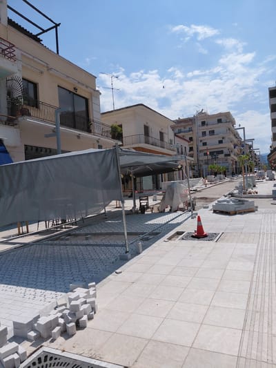 This is a medium-distance, eye-level, outdoor, daytime photograph of a street under construction in Aigio, Greece. The scene is dominated by a street undergoing paving work, with stacks of light-colored paving stones visible in the foreground and scattered along the sides of the road. Construction barriers, including a large gray tarp supported by metal poles, partially obscure the view of a building on the left. The street is lined with multi-story buildings, some with balconies and awnings. Several streetlights and trees are also present. An orange traffic cone sits on a manhole cover in the foreground. The sky is blue with scattered white clouds. The overall impression is one of ongoing urban development and renovation.