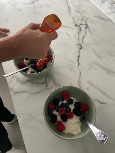 Overhead shot of a marble countertop on which there are two light green bowls filled with yogurt and berries (raspberries, blueberries, blackberries). The bowl on the left is having honey poured over it from a clear, orange-tinted container held by a person's left hand. A silver spoon is in each bowl. A person is standing at the counter wearing black pants and white socks. The location is likely a private residence in Welwyn Hatfield, United Kingdom, based on the provided location data.