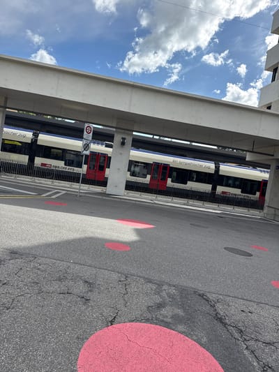 The image captures an oblique view of a train station or platform area in Liestal, Switzerland, under a bright, partly cloudy sky. A white and blue Swiss Federal Railways (SBB CFF FFS) passenger train, featuring red doors and the text "ein sympathischer zug." (a friendly train) on its side, is stopped at a platform.
Above the train and dominating the upper left, a large, light-grey concrete structure, likely an overhead bridge or part of the station's architecture, extends across the frame. The ground in the foreground is dark grey asphalt, showing some cracks, and is marked with several large, vibrant pink/red circles and a smaller dark grey circle. A black metal fence separates the train tracks from this asphalt area.
A white circular road sign with a red border and a horizontal black bar, indicating a "No entry" restriction, is visible to the left of the train, mounted on a pole. Below it, a supplementary sign reads "ZONE". The sky is clear blue with scattered white cumulus clouds, indicating a sunny day, likely morning or afternoon, with shadows visible on the ground and train. No people are depicted, highlighting the infrastructure and the train as the main subjects.