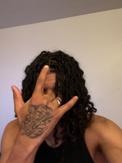 An angled close-up shot of a person with dark, shoulder-length dreadlocks. The person is wearing a black tank top. Their hand is raised, obscuring the upper part of their face, with the fingers forming a "devil horns" gesture. A silver ring is visible on one of the fingers. The hand has a large, ornate tattoo in black ink. The background is a plain, light-colored wall. There is nothing in the image to indicate the city of Massy, France.