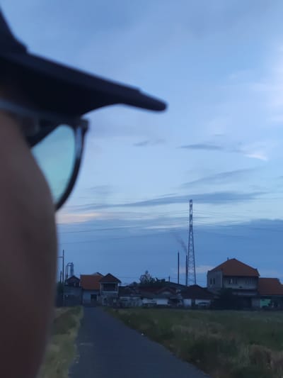 The image is an eye-level, outdoor shot of a street in Banjarkemantren, Indonesia, at dusk. A person wearing sunglasses is in the foreground, with their face blurred and partially obscured by their hand. The sunglasses lens reflects the sky and some of the landscape.

The background shows a residential area with several houses, some of which appear to be two-story structures with tiled roofs. A paved road leads into the distance, with grassy areas on either side. Further in the distance, a tall, lattice-style transmission tower is visible, with power lines stretching across the sky. There is a faint plume of smoke emanating from a structure behind the transmission tower. The sky is a gradient of blue and gray, with some wispy clouds, suggesting the end of daylight. The overall atmosphere is serene and rural.