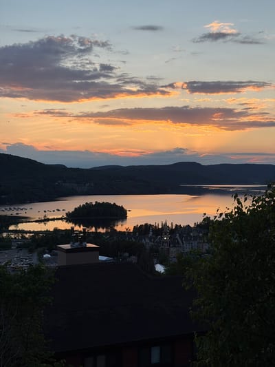 The image depicts a scenic view of Mont-Tremblant, Quebec, Canada, at dusk. The foreground is dominated by the dark roof of a building, with trees on the right partially obscuring the view. Below, a small cluster of buildings with steeply pitched roofs and a clock tower is visible, suggesting a village or resort setting. Further in the midground, a body of water, likely a lake, reflects the warm hues of the sunset. Several boats are moored on the left side of the lake, and a small, tree-covered island is situated in the water. The background features rolling, tree-covered hills silhouetted against a sky filled with a gradient of colors from pale blue at the top to vibrant orange and pink near the horizon, characteristic of a sunset. Scattered clouds add texture to the sky. The overall atmosphere is serene and picturesque, highlighting the natural beauty of the area.