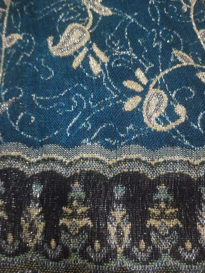 A close-up shot reveals intricate textile details. The upper portion features a teal fabric with silver and beige floral or paisley patterns. A wavy, horizontally oriented border separates this section from the lower part, which is predominantly black with similar floral or abstract designs highlighted by the same silver and beige threads. The overall impression is that of an ornate and potentially luxurious fabric.