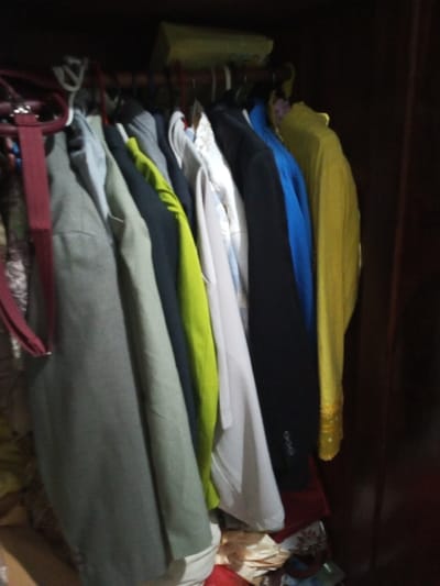 A closet's interior reveals clothes hanging on a rod, with a yellow box above. Garments in various colors are visible: shades of gray, green, white, black, blue, and yellow. The lower portion is cluttered with additional clothing and items, partially obscured. Dark wood panels of the closet frame the scene.