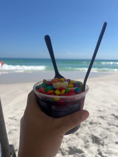 The image displays a first-person perspective, showing a hand holding a cup of dessert against a vibrant beach backdrop. The image is oriented sideways, with the horizon running vertically.
In the foreground, a light-skinned hand firmly grips a clear plastic cup on its right side. Inside the cup, there's a colorful dessert, likely a frozen treat such as açaí. The top of the dessert is generously covered with various toppings: numerous small, round, candy-coated chocolates in multiple bright colors (red, blue, yellow, green, orange, brown), and several slices of banana arranged centrally. Some red fruit pieces, possibly strawberries, are also visible among the candy and bananas. Two black plastic utensils, one appearing to be a long spoon or stirrer and the other a shorter spoon, are inserted into the dessert. On the side of the cup, there's a dark label with small, indistinct white text, which is unreadable due to resolution and size.
The background reveals a bright, sunny beach scene. To the left, a large expanse of clear, bright blue sky is visible. Below the sky and to the right of the hand holding the cup, there's the ocean, with light blue and turquoise waters indicating a shallow or clear area. Gentle waves can be seen breaking on a light-colored sandy beach, which occupies the right side of the background. In the distance, near the water's edge, a small red flag is visible.
The overall scene suggests a leisurely moment, enjoying a refreshing treat by the sea on a clear, warm day.